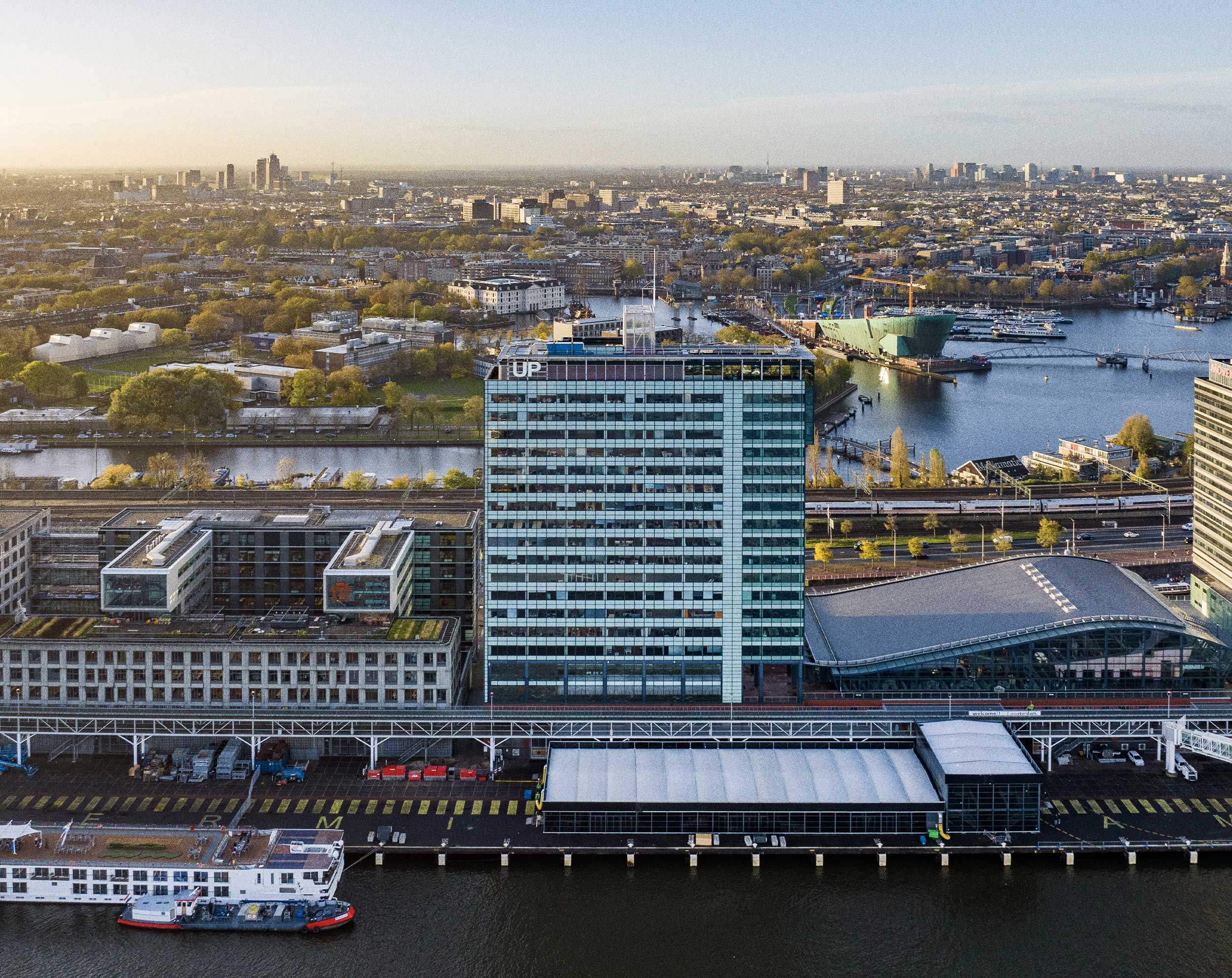 Luchtfoto van een kantoorgebouw aan het water met een “UP”-logo, omringd door kanalen, wegen en de stadsskyline.