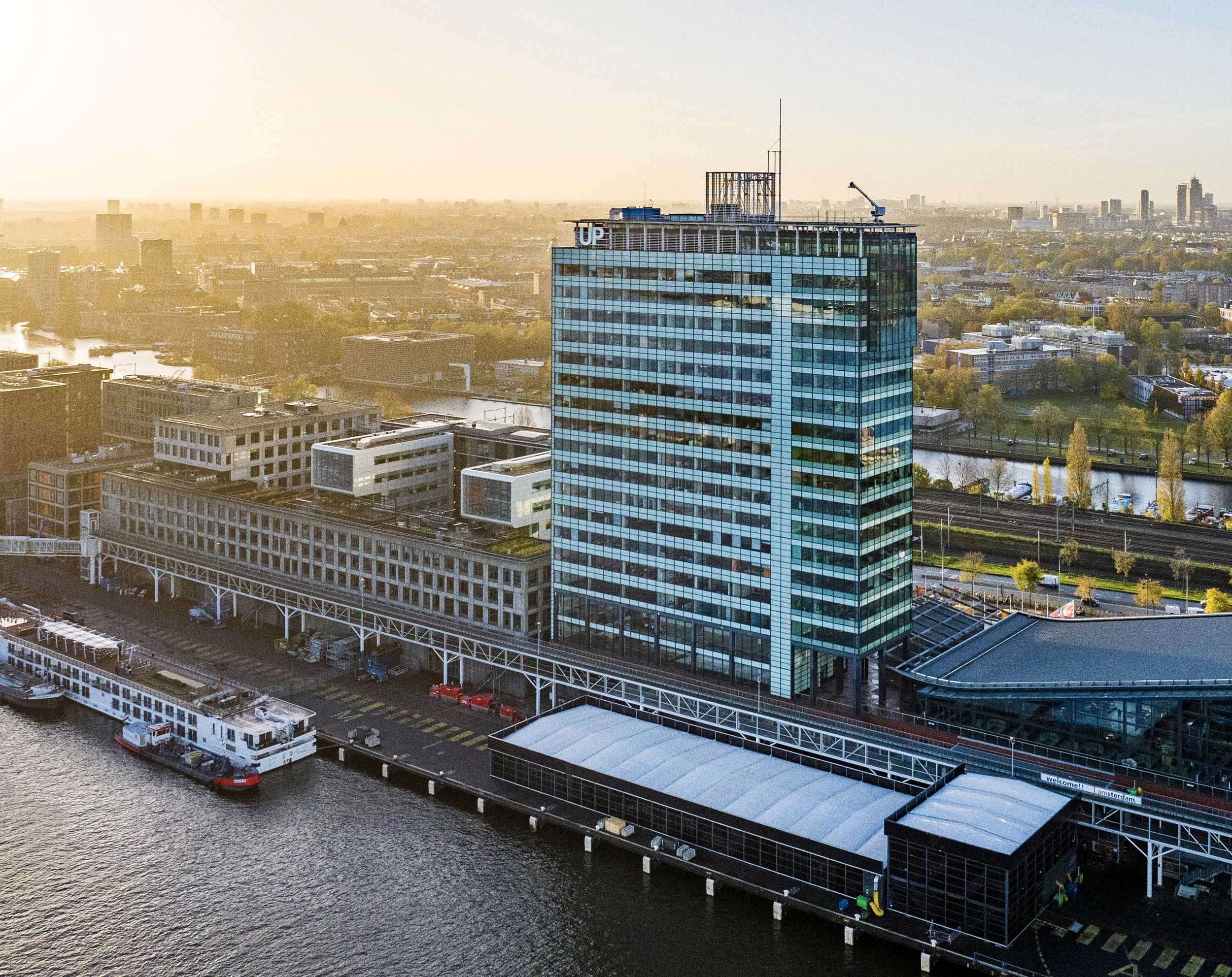 Luchtfoto van een hoog gebouw aan het water met een groot “UP”-logo, verlicht door warm avondlicht