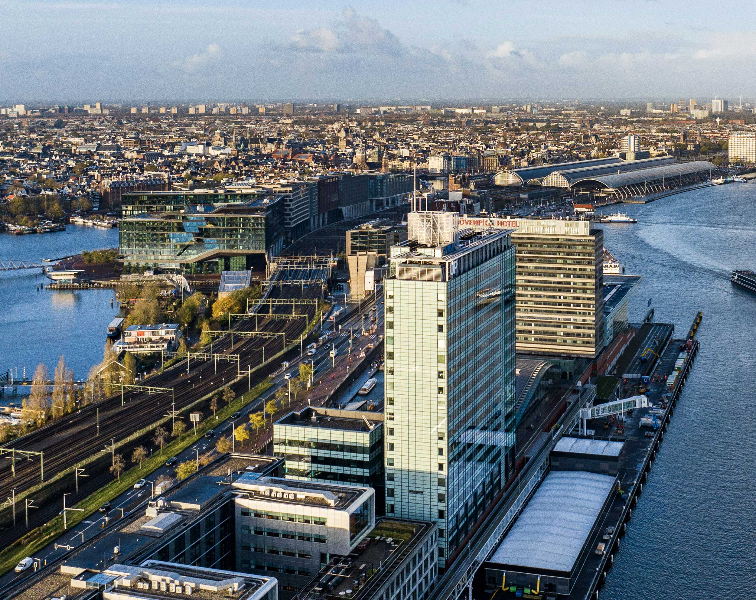 Luchtfoto van de Amsterdamse waterfront met moderne gebouwen, spoorlijnen en het water.