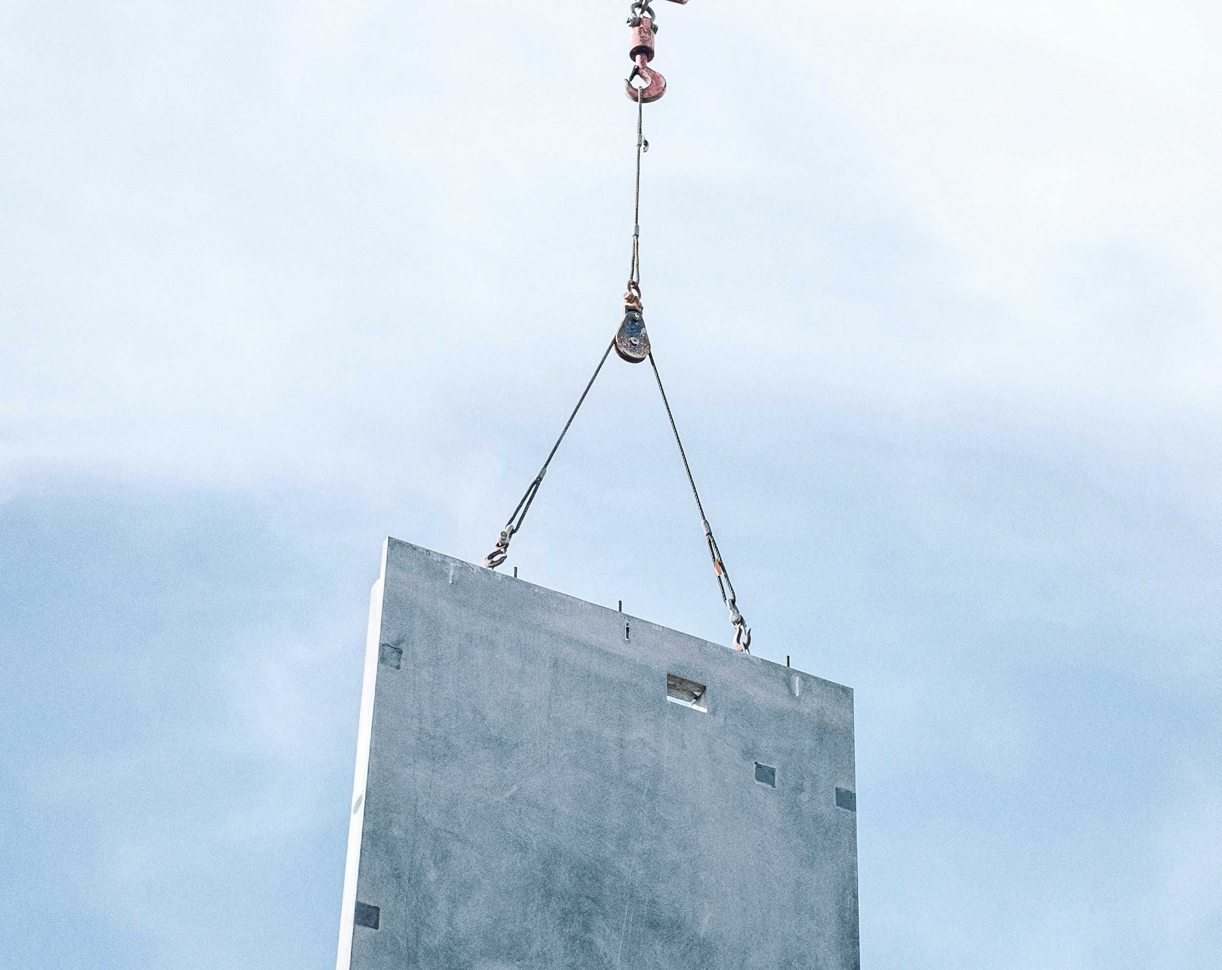 Betonnen wandpaneel dat met kraankabels omhoog wordt gehesen tegen een lichte lucht.