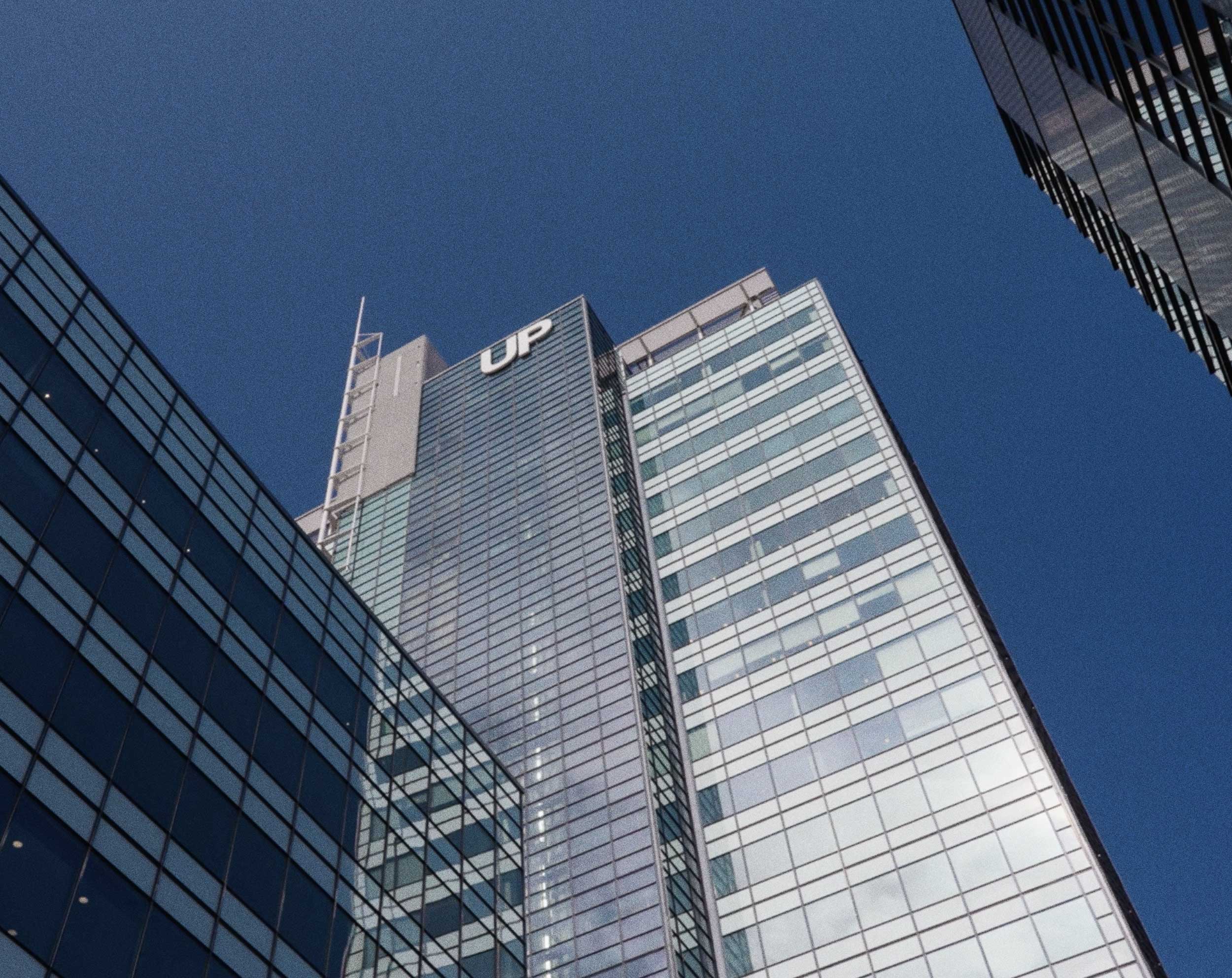Laag perspectief op een moderne glazen hoogbouw met een groot “UP”-logo tegen een heldere blauwe lucht.